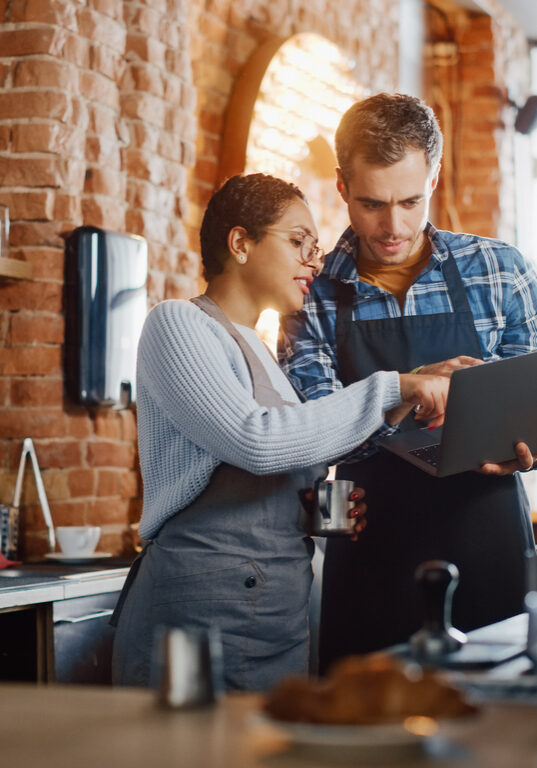 Two Diverse Entrepreneurs Have a Team Meeting in Their Stylish Coffee Shop. Barista and Cafe Owner Discuss Work Schedule and Menu on Laptop Computer. Multiethnic Female and Male Restaurant Employees.